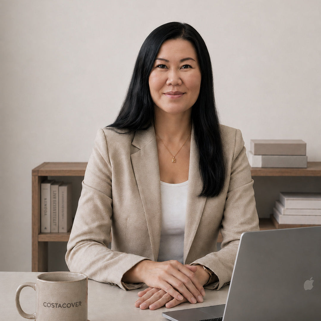 A woman with long black hair sits at a desk wearing a beige blazer over a white top. In front of her is an open silver laptop