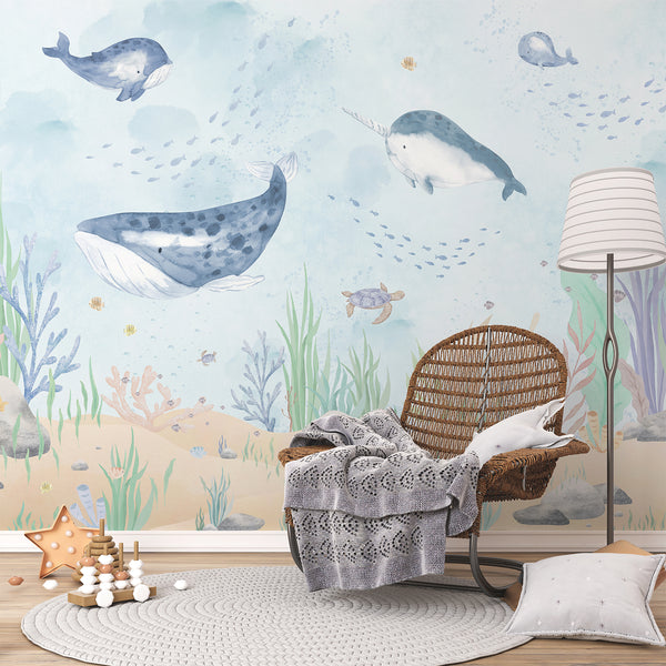 Ocean nursery wallpaper with large blue whales, a narwhal, sea turtle, fish schools, bubbles, coral, and seaweed on a pale blue background.