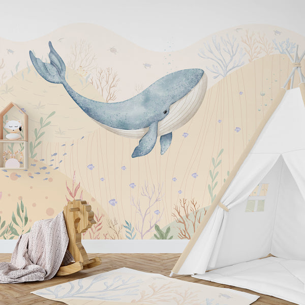 Watercolor blue whale wallpaper featuring a large whale, tiny fish, bubbles, coral, seaweed, and soft sandy ocean hills in pastel coastal colors.