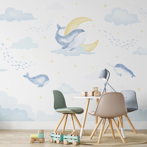 Blue ocean nursery wallpaper with whimsical watercolor whales swimming among clouds, tiny fish, crescent moons and yellow stars for a baby room.