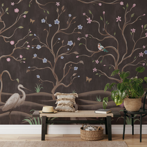 Vintage chinoiserie wall mural with white herons, pink and blue flowers, twisting branches and birds against a rich dark brown background.