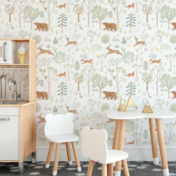 A children's playroom corner featuring a wall covered in whimsical forest-themed wallpaper with bears, deer, trees, and foxes in muted earthy tones. In the foreground, there is a small white table and two matching chairs.