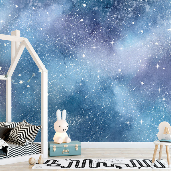 A children's bedroom scene featuring a large wall mural with a blue and black starry galaxy pattern. The room includes a white house-frame bed, a small white chair, a black-and-white striped pillow.
