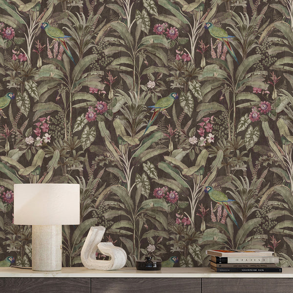 Dark tropical wallpaper featuring green parrots, muted pink flowers and lush botanical foliage on a deep brown background with a vintage jungle look.
